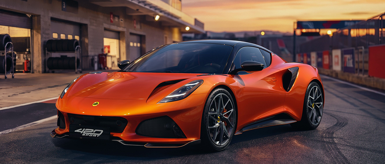 alt="Lotus Emira 420 Sport orange métallisé sur la ligne des stands d'un circuit au coucher du soleil"