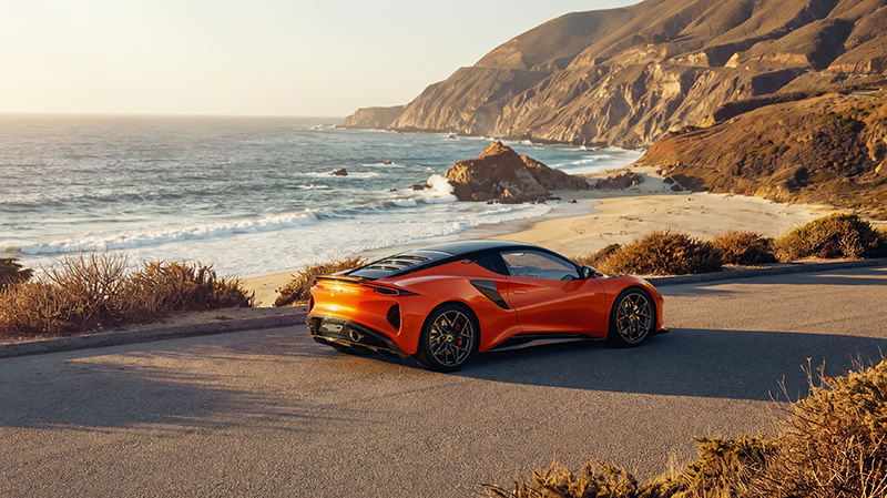 alt="Vue arrière de trois quarts d'une Lotus Emira 420 Sport orange métallisé sur un circuit automobile au crépuscule, mettant en valeur le hayon à persiennes et le diffuseur arrière"