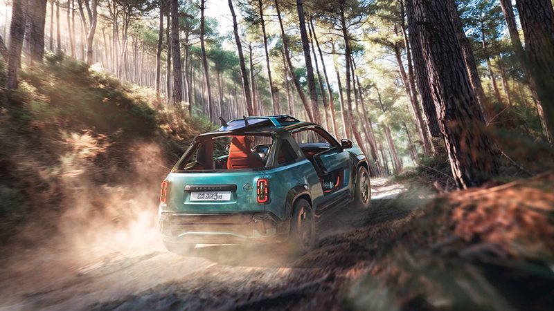 Vue arrière d'une Renault 4 JP4x4 Concept Vert Émeraude avec planche de surf sur le toit, roulant en tout-terrain sur un sentier poussiéreux dans une forêt de pins.