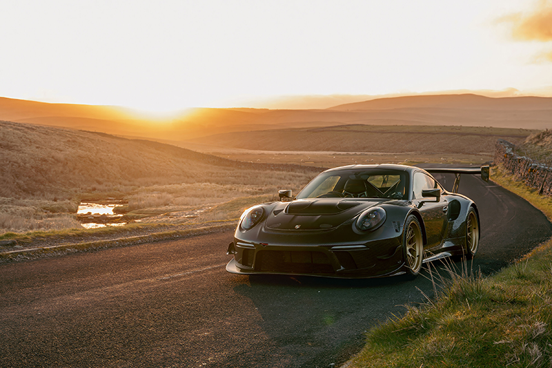 TaPorsche 911 RT par Talos Vehicles en fibre de carbone apparente avec jantes dorées, photographiée de face sur une route de montagne au coucher du soleil.