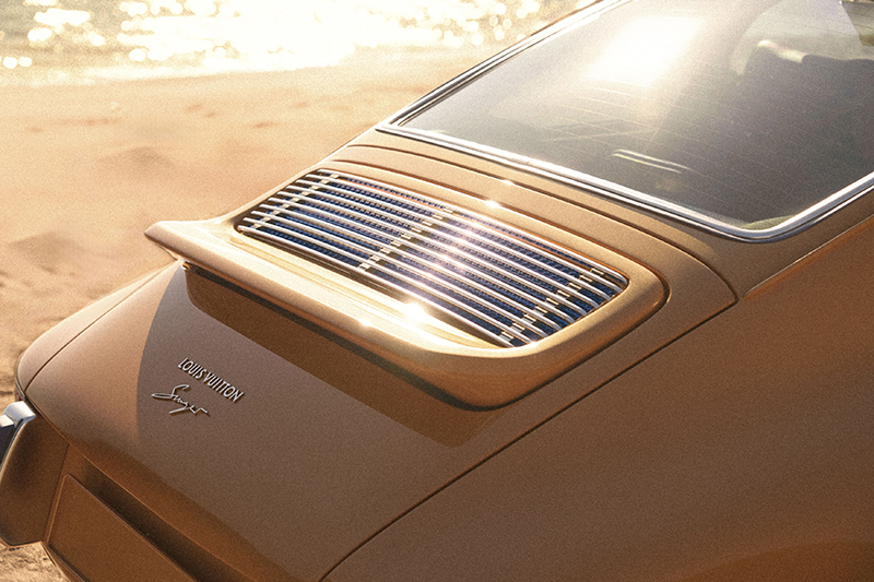 Gros plan sur le capot moteur d'une Porsche 911 Singer Classic orange safran, montrant la grille d'aération brillante, le reflet du soleil et le logo "LOUIS VUITTON X Singer" en métal argenté.