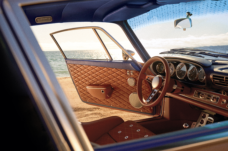 Vue intérieure d'une Porsche 911 Singer garée face à la mer, montrant la portière ouverte garnie de cuir marron matelassé et le tableau de bord bleu.